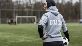 Jonge vrouwelijke voetbalcoach met hoofddoek op aan de rand van een voetbalveld