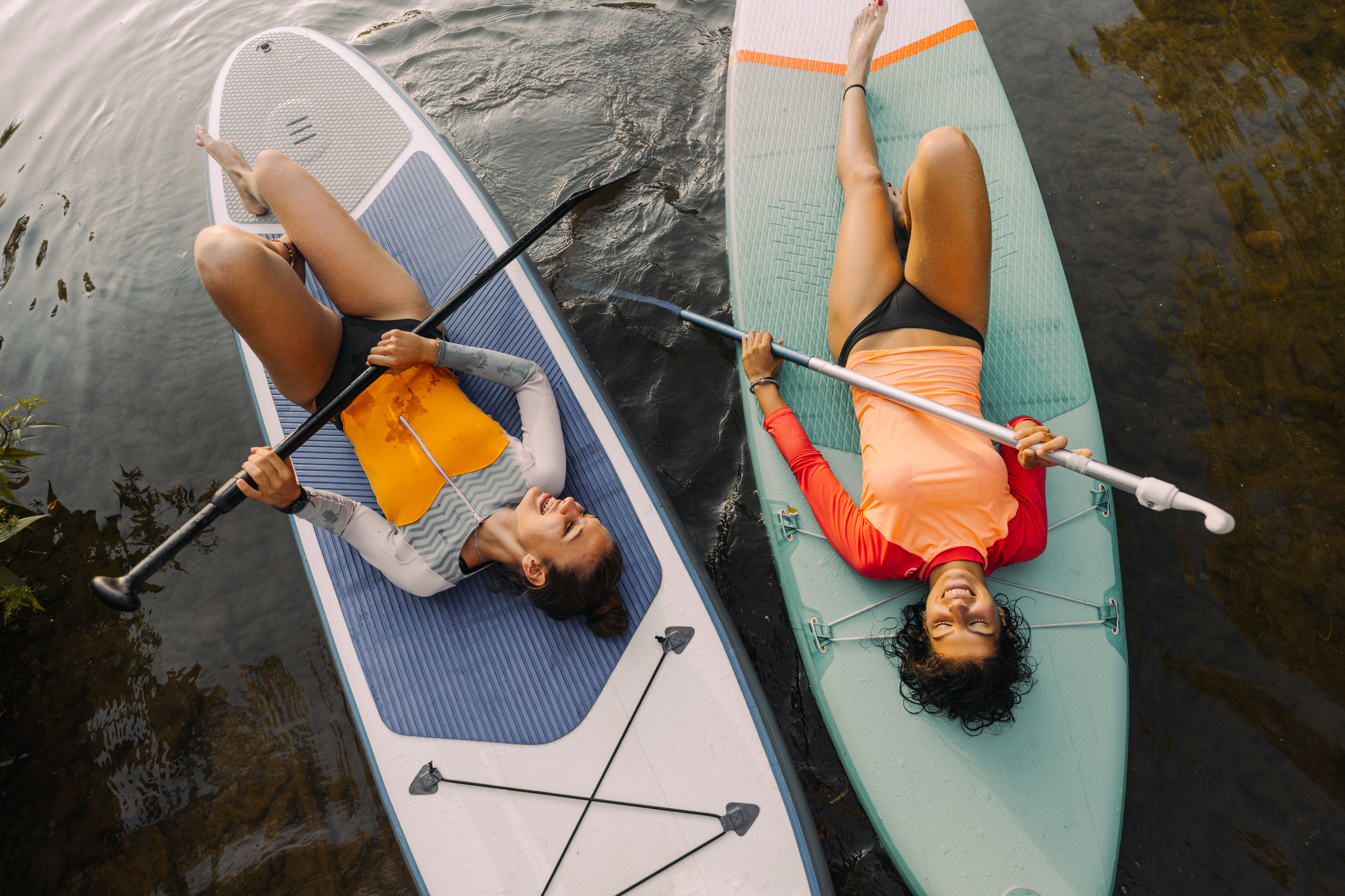 Photo of two teenage girls lying on their stand-up paddleboards and having a great time; teenagers hanging out in nature and enjoying their summer vacation.