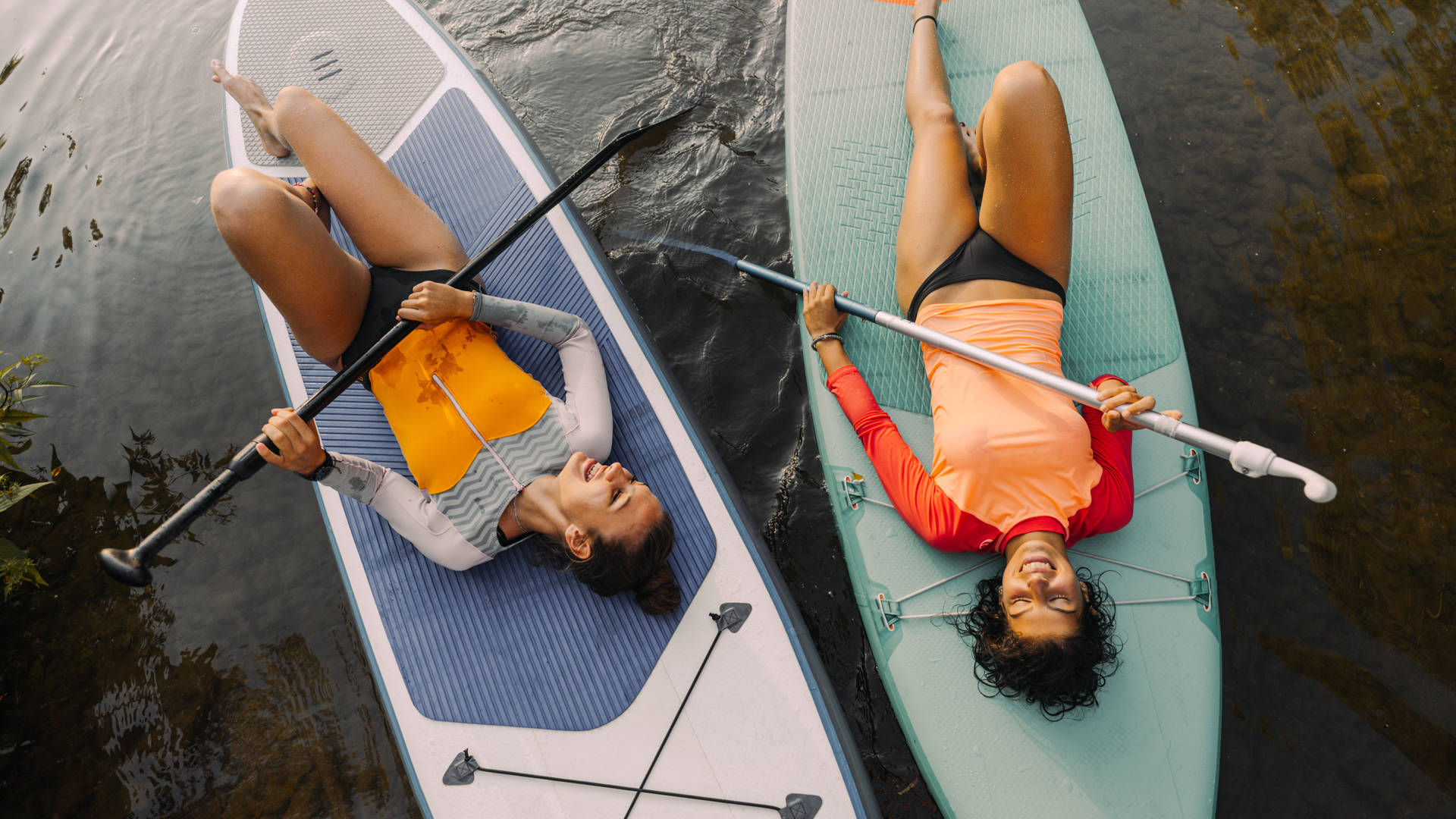 Photo of two teenage girls lying on their stand-up paddleboards and having a great time; teenagers hanging out in nature and enjoying their summer vacation.