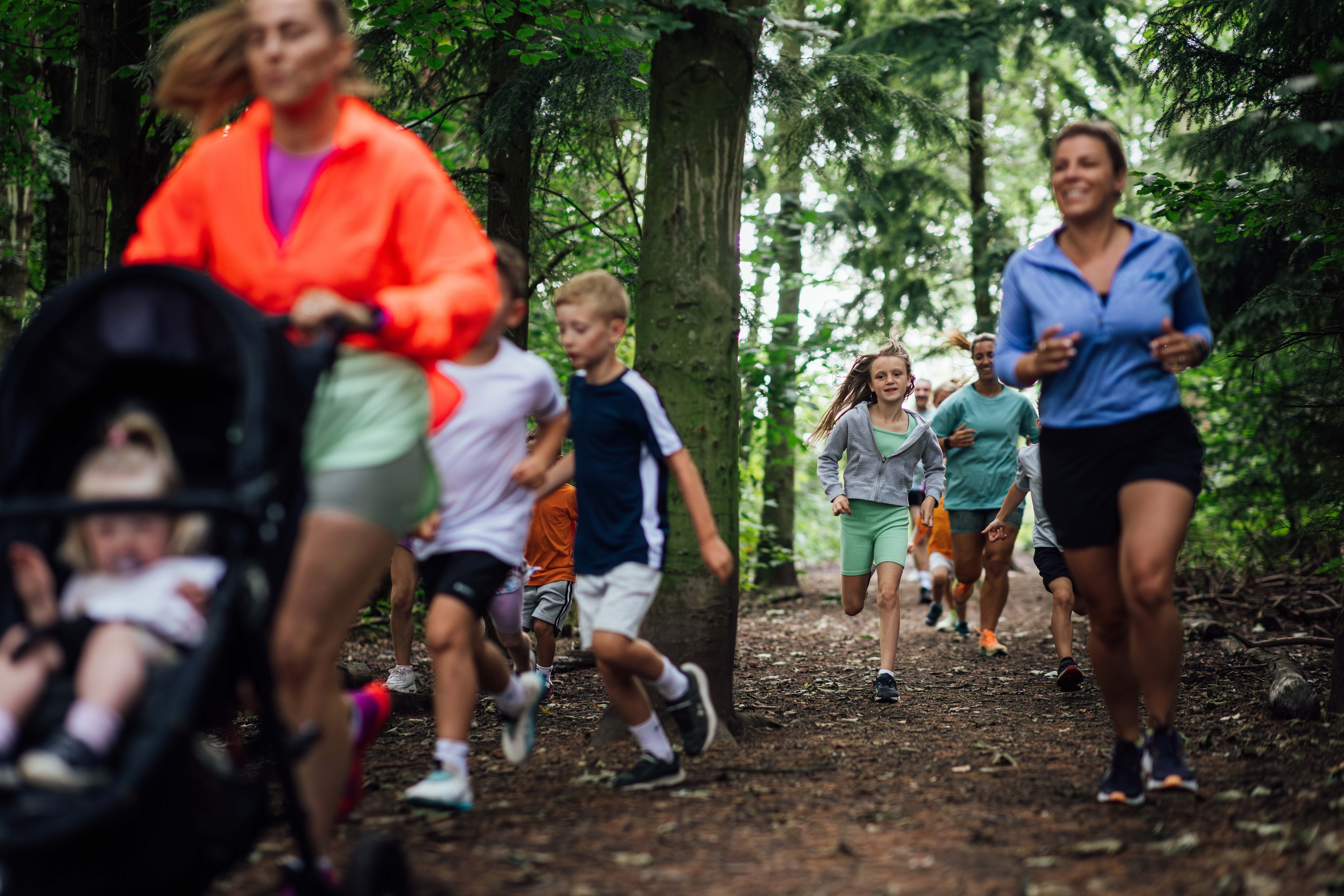 A medium shot of a group of adults and children wearing sports clothing. They are taking part in a fun run with other children and adults at Cramlington Nature Reserve in the North East of England. The focus is on a young girl as she runs with other participants.