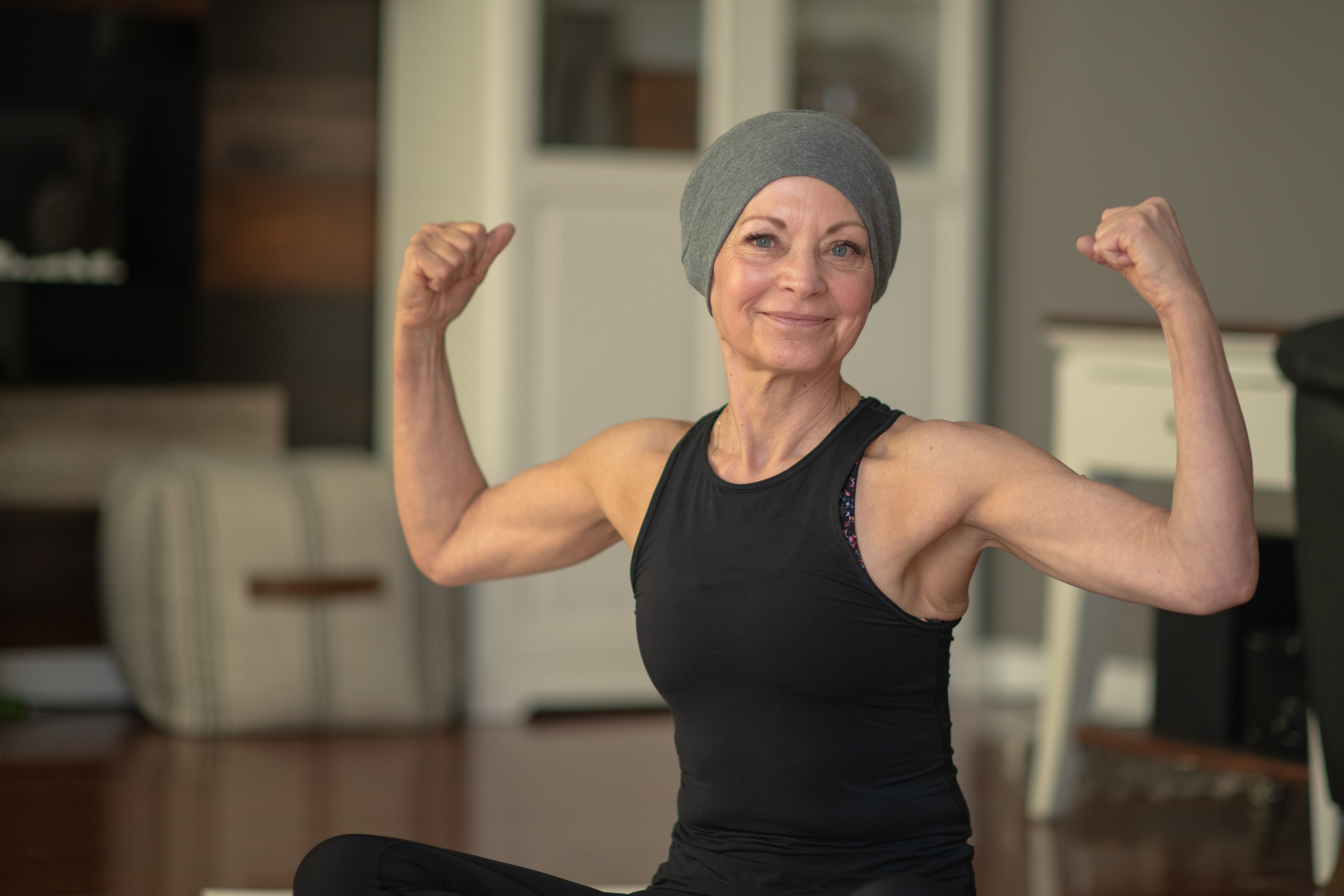 A senior woman with cancer flexes her muscles while sitting at home on her floor. She is smiling because she is getting stronger.