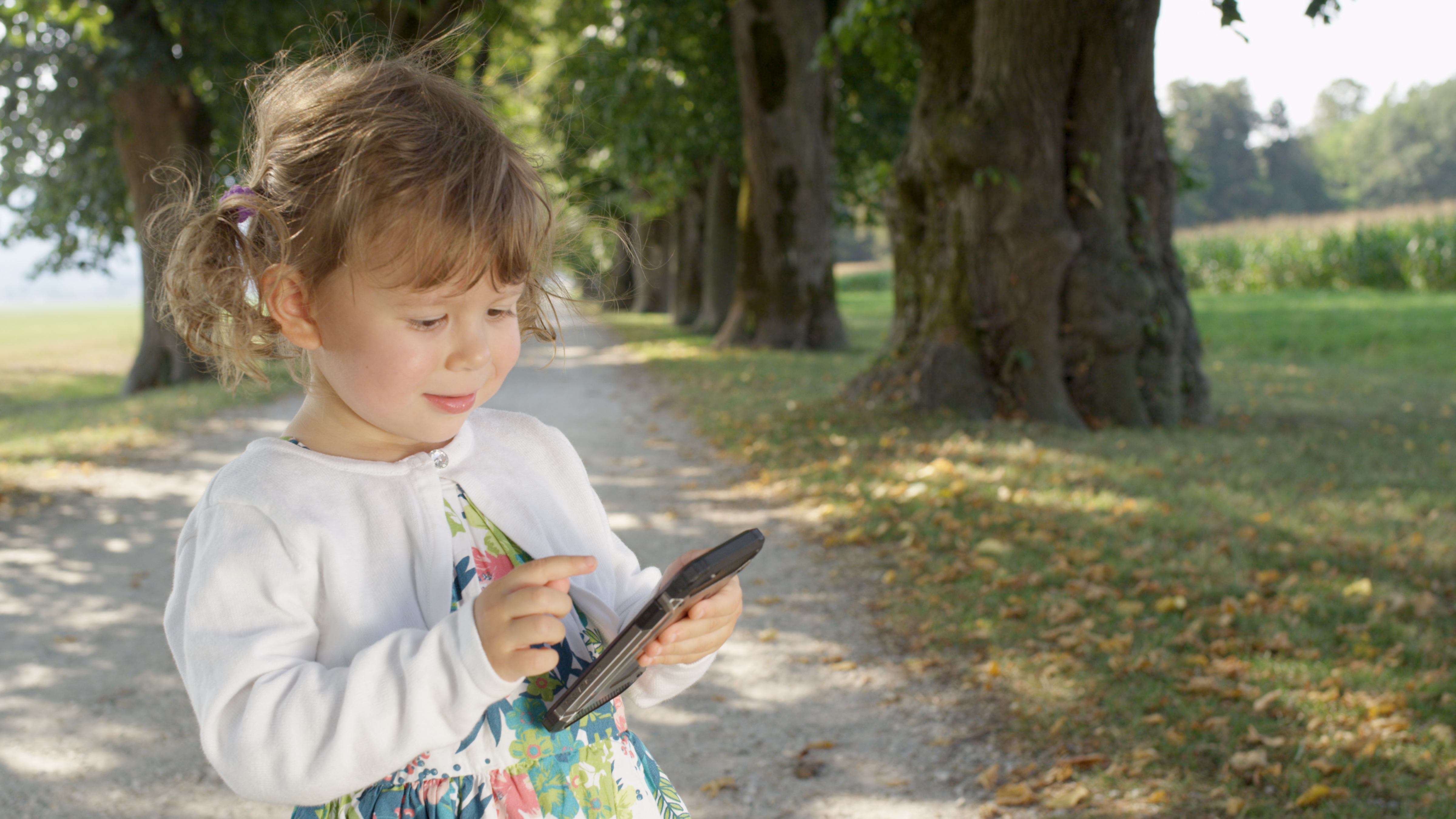 CLOSE UP: Adorable young girl types with her tiny fingers on a modern mobile phone. Cute baby daughter playing games at the sunny park on her father's cell phone. Toddler holding a cool smart phone.