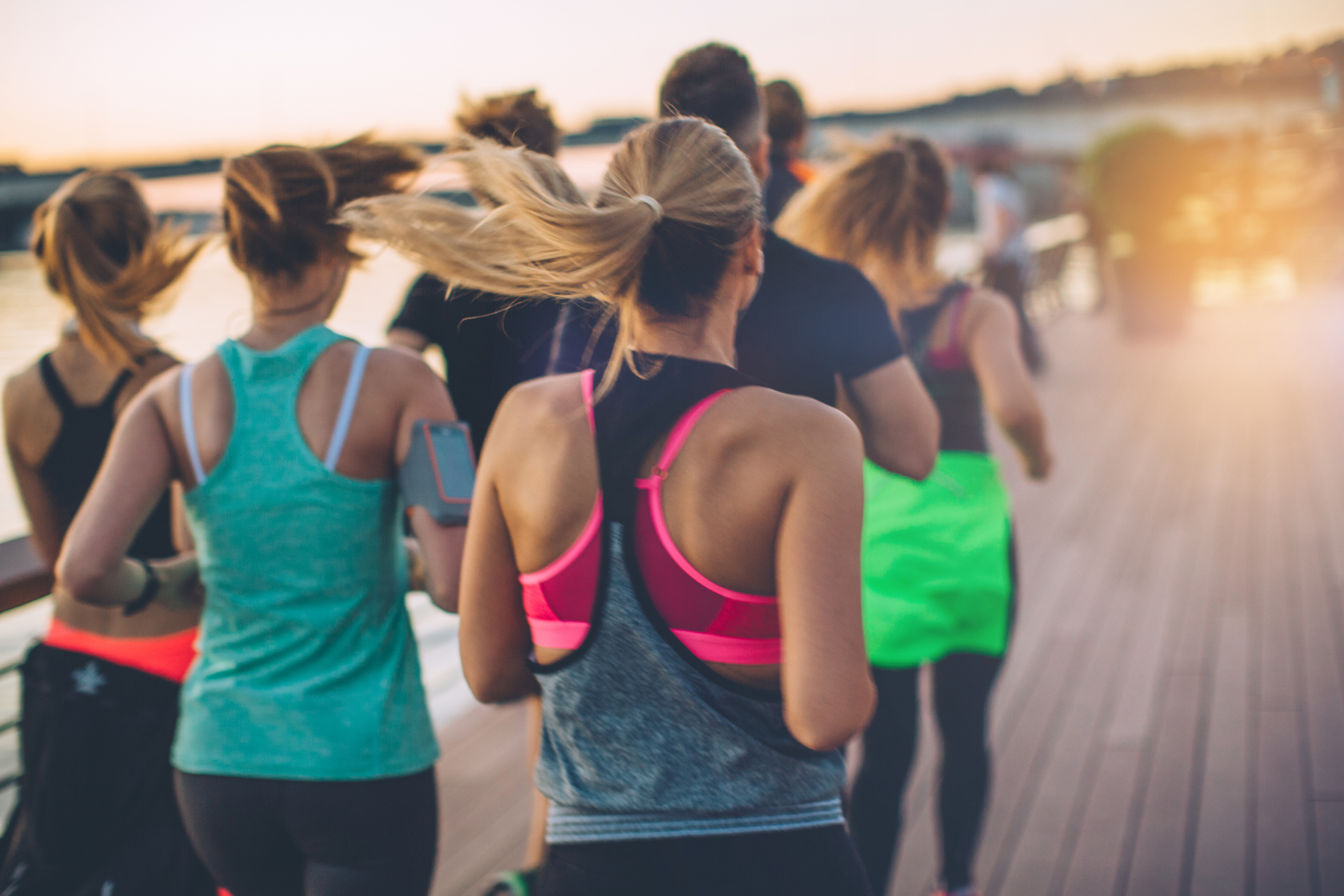 Group of young people competing in a race. Young men and women on riverside promenade running. They are wearing sport clothing. View from behind.