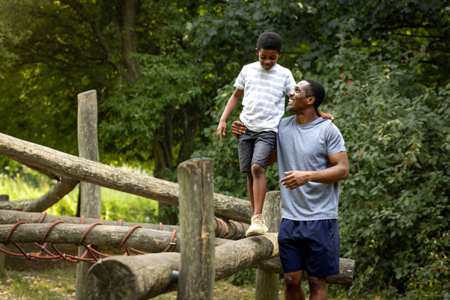 A father and son sending quality time together in their local park. The young boy is balancing on a obsticle course while his dad supports him by the waist. the father is looking up at his son with a smile.