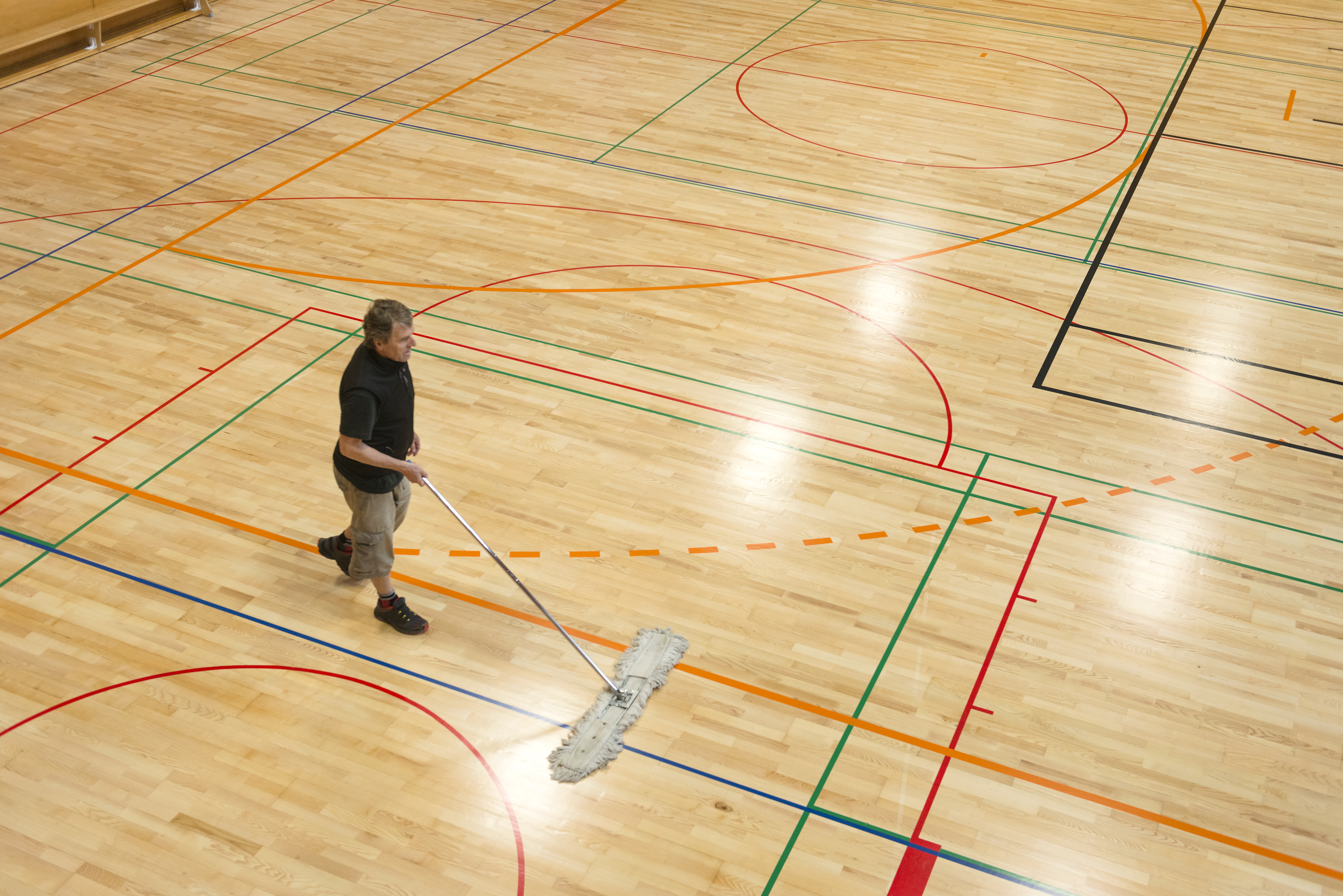Senior man in shorts and black T-shirt cleaning the floor in a new school gymnasium, new parquet with lines and curves for different team sports(basketball, volleyball, soccer, handball). Side view from above, daylight from roof windows. Nikon D800, full frame, XXL. Motion blur.
