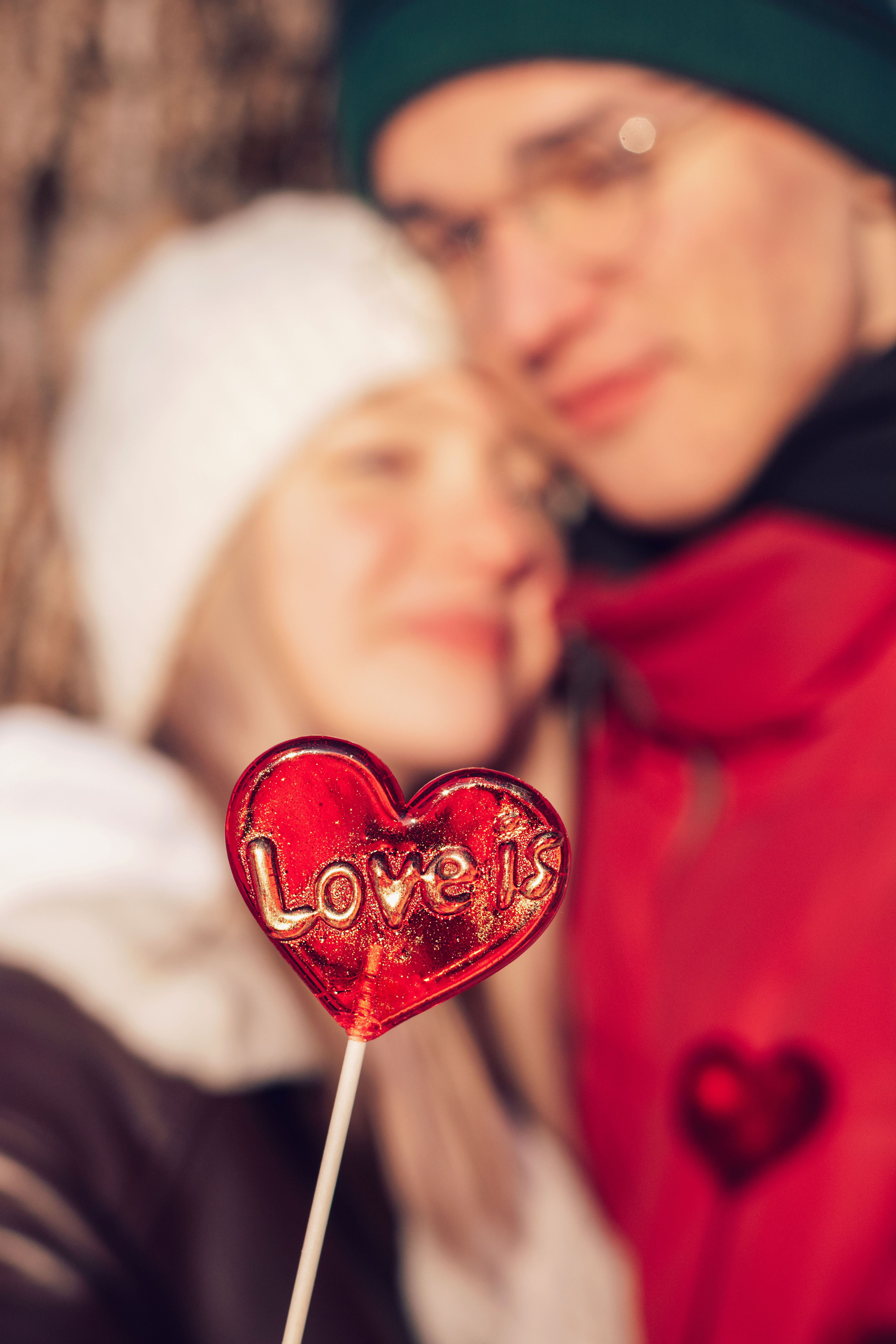 Blurred young couple in love on a date on a winter day.In the foreground is a red lollipop in the shape of a heart and the inscription Love is.Valentine's day concept.