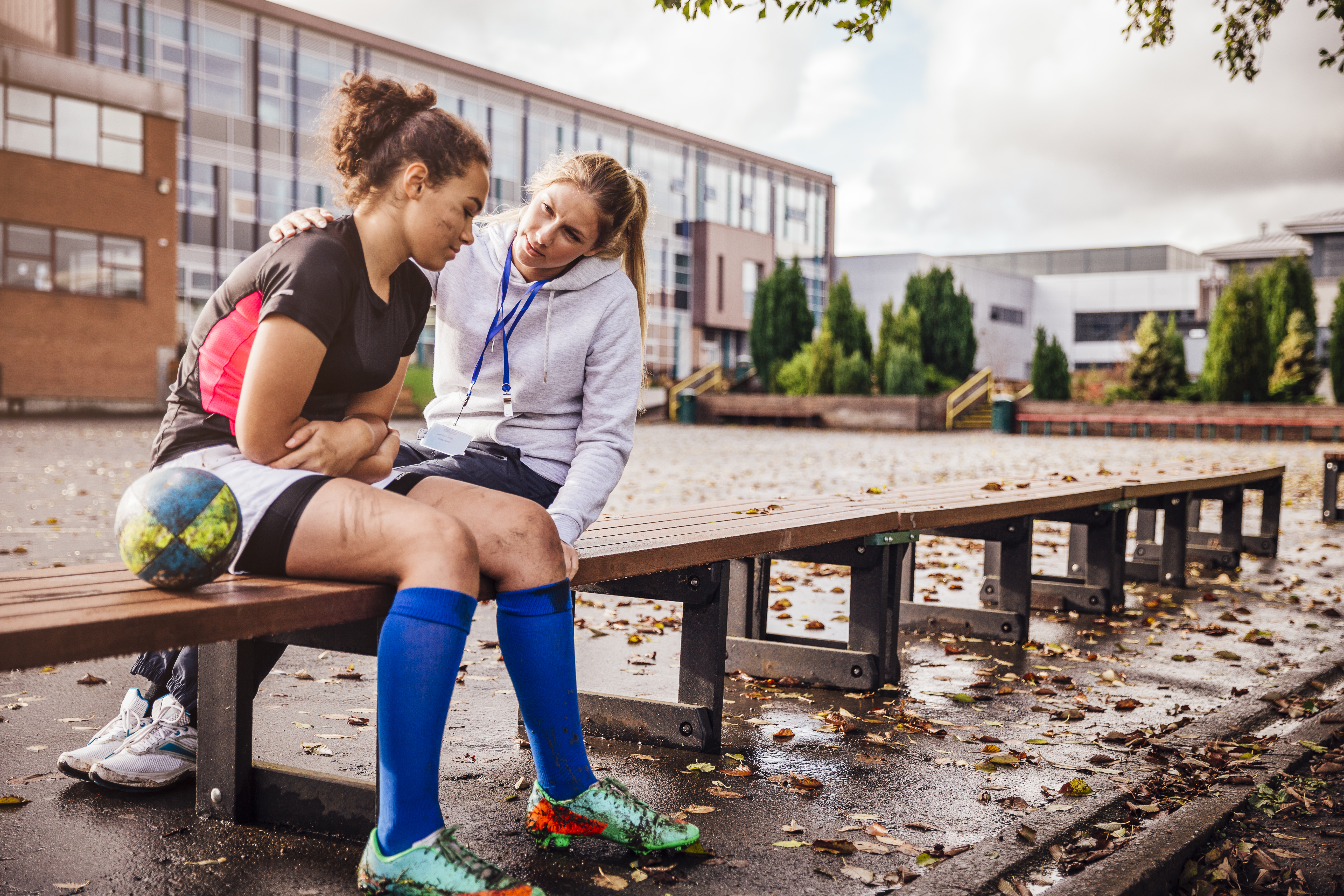Teenage girl sitting on a bench with her rugby ball at one side. Dirty with mud after her match. Female coach sits close supporting her as the girl looks sad.