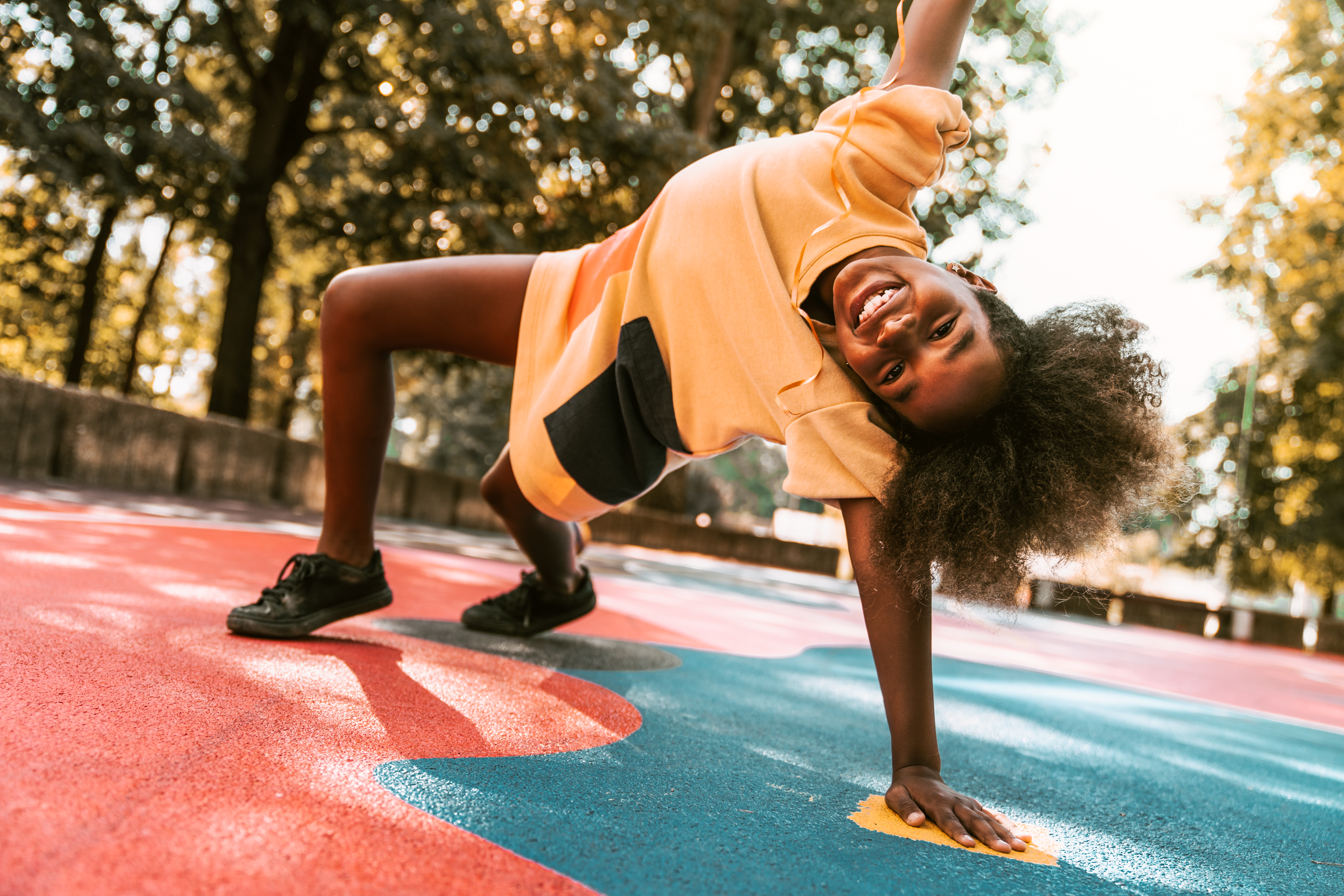 A young girl is playing around the basketball court. She is stretching on one arm, the other is high in the air. She is happy to be outside on a warm summer day.